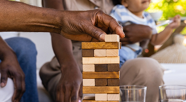 A person places a block on a tabletop tower game while family members sit together in the background.